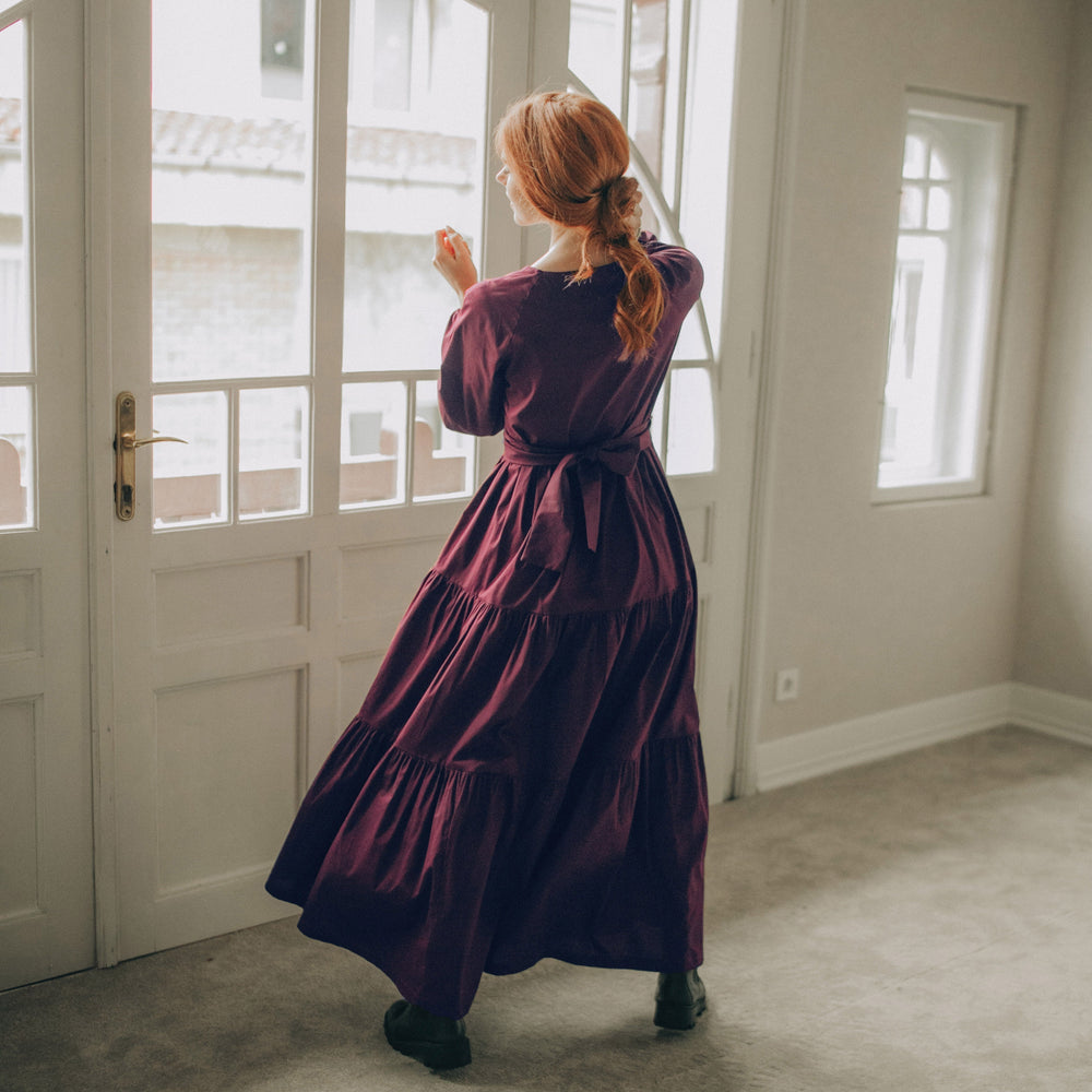 
                      
                        Woman in a long purple dress standing in a room with large windows.
                      
                    