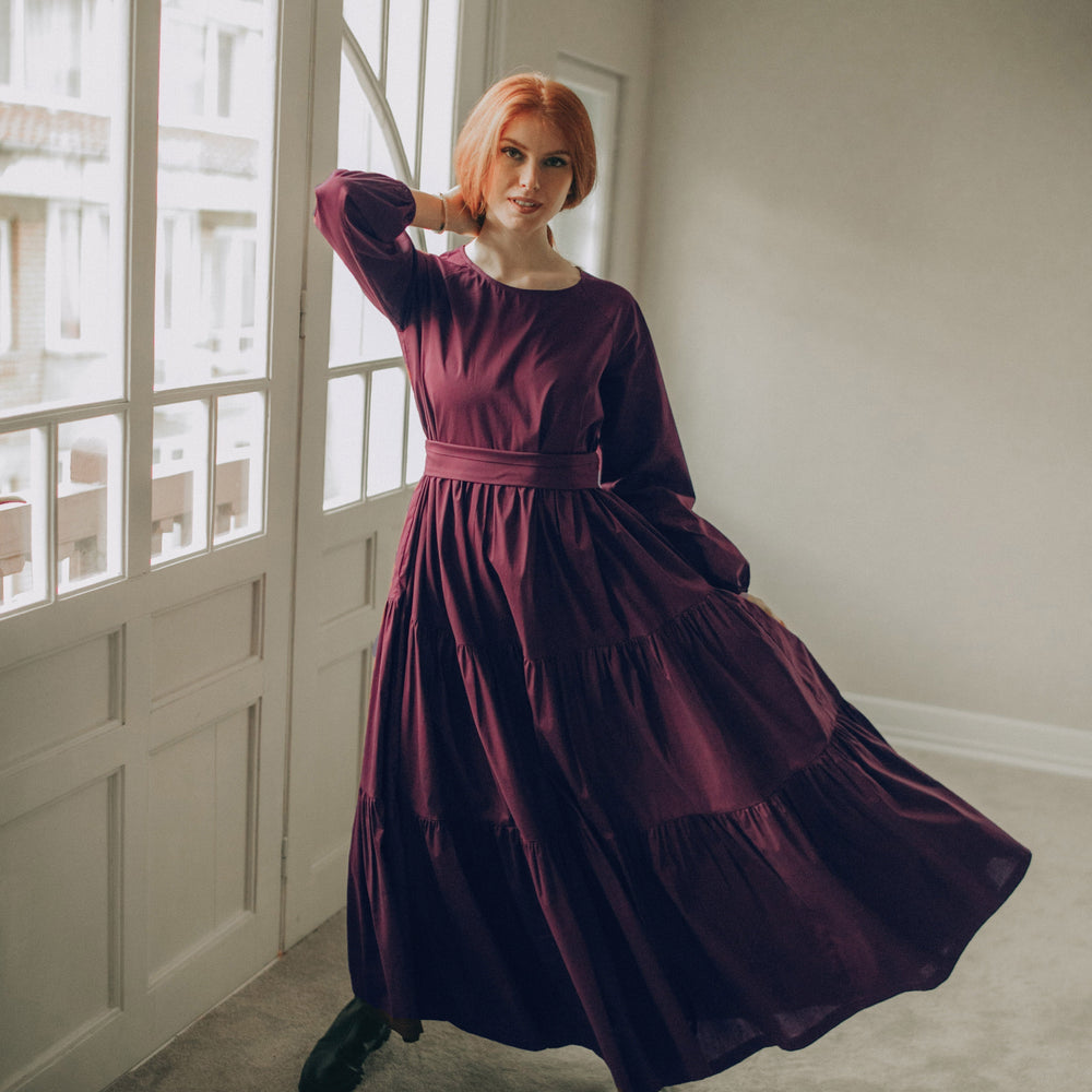
                      
                        Woman in a long burgundy dress standing in a room with large windows.
                      
                    