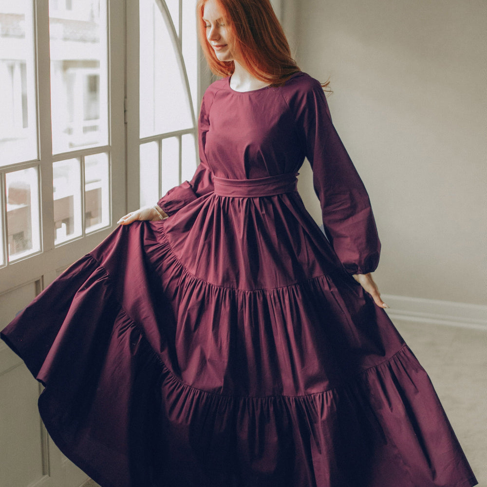 Woman wearing a deep burgundy tiered cotton maxi dress with long sleeves and adjustable tie waist standing by a window