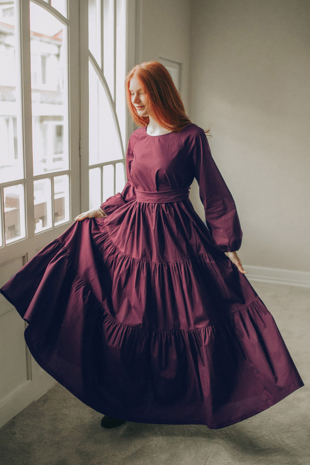 Woman wearing a deep burgundy tiered cotton maxi dress with long sleeves and adjustable tie waist standing by a window