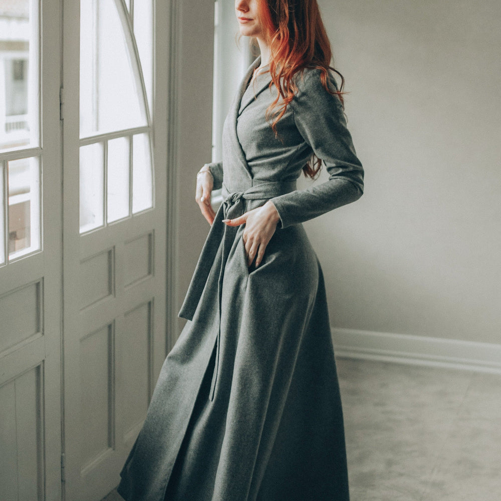 Woman wearing long gray wool wrap dress with shawl collar and side pockets indoors near window