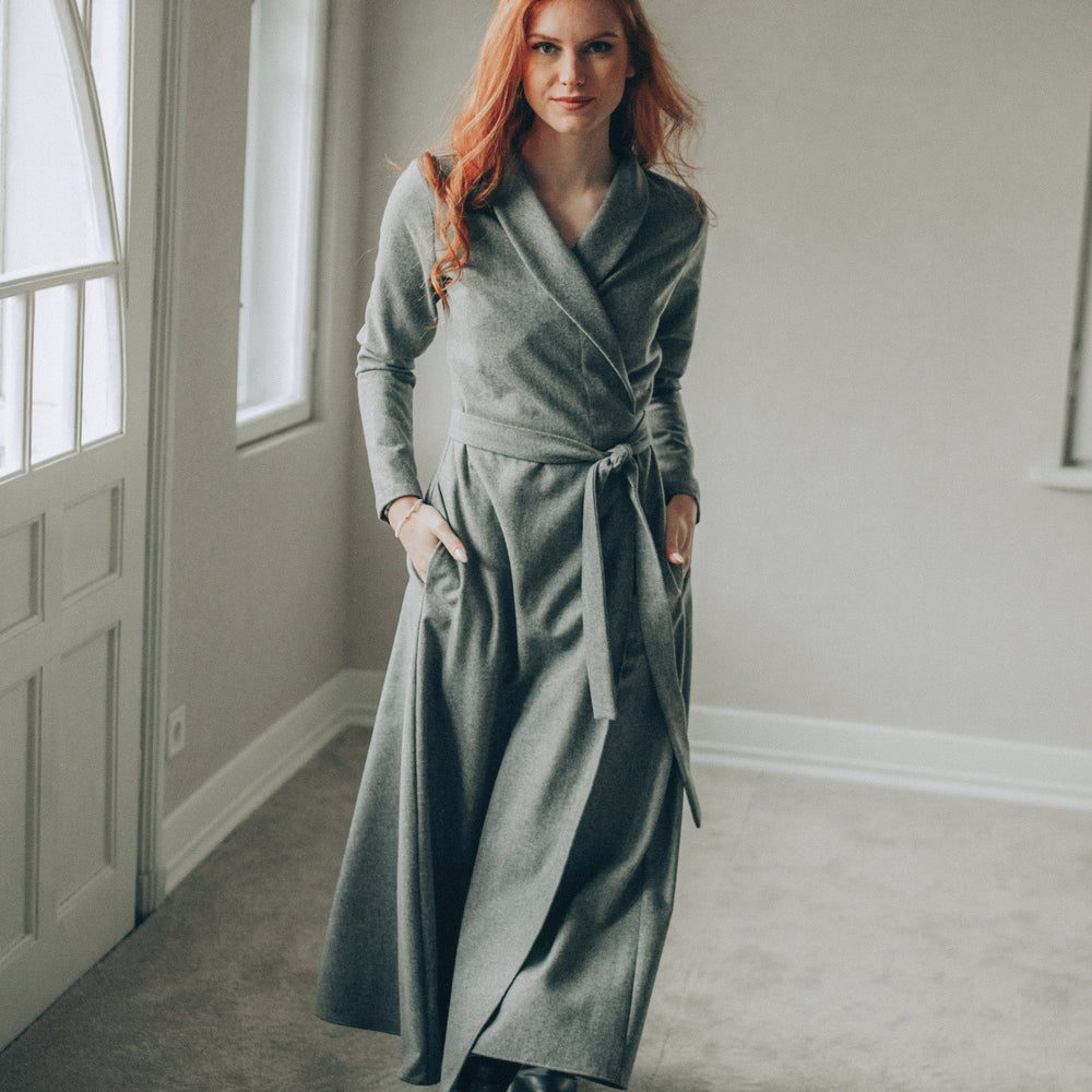 Woman in a long gray dress standing in a room with large windows.