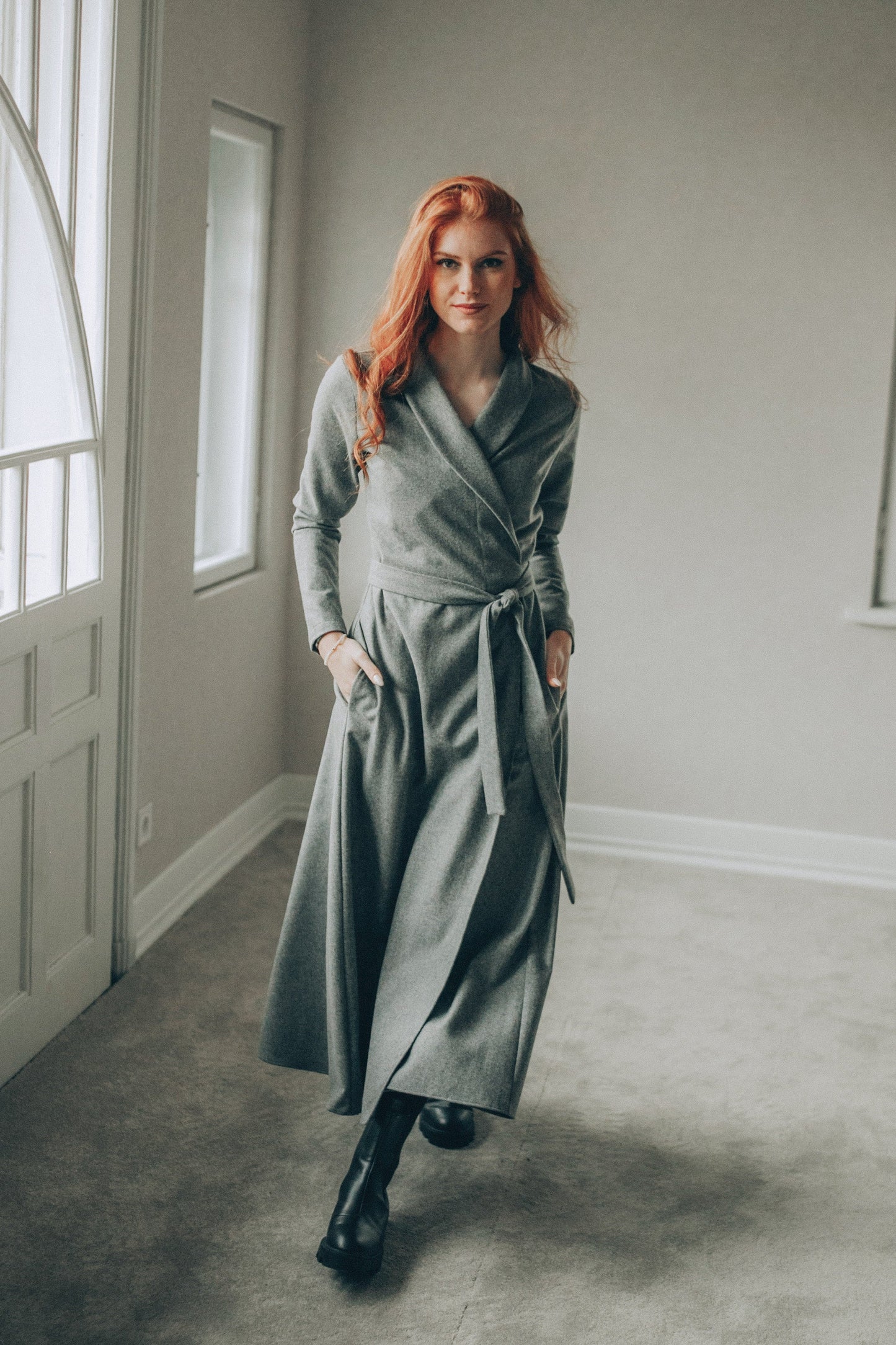 Woman in a long gray dress standing in a room with large windows.