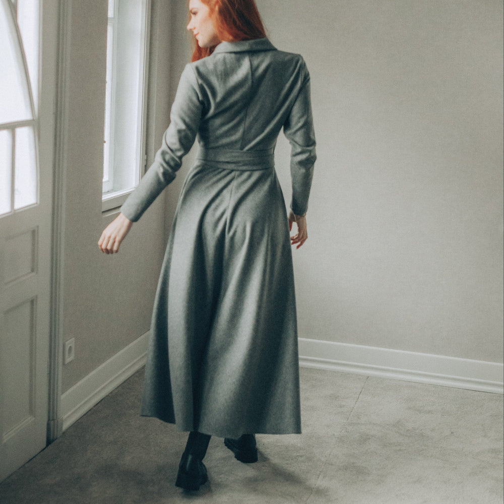 
                      
                        Woman in a long gray dress standing in a room with a window.
                      
                    