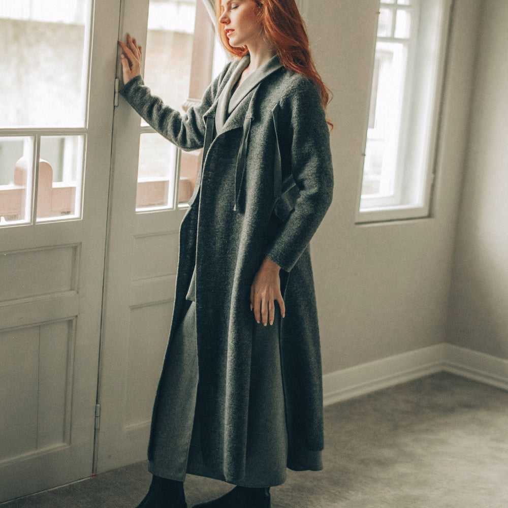 Woman in a long cardigan standing in a room with large windows.