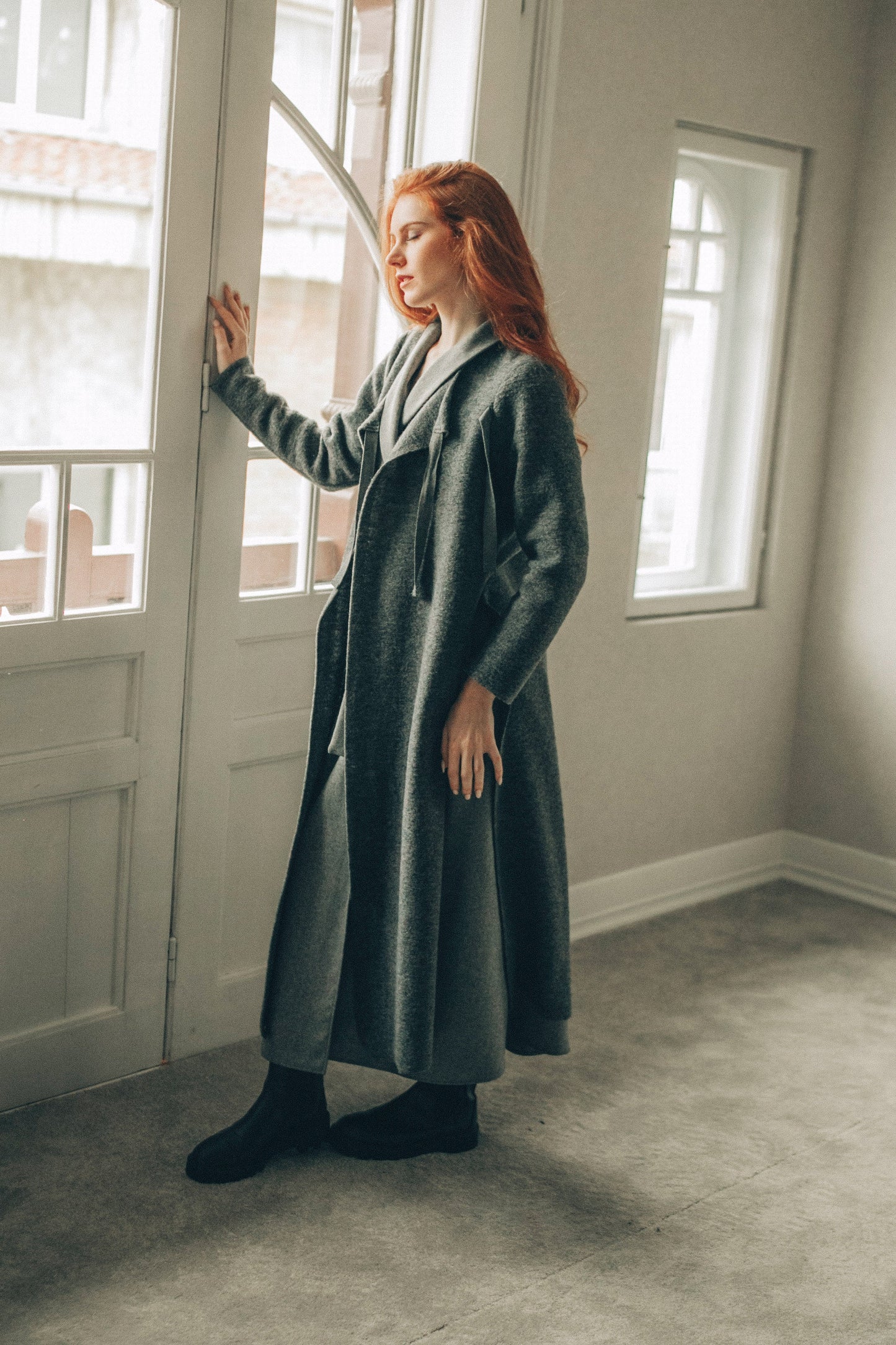 Woman in a long cardigan standing in a room with large windows.
