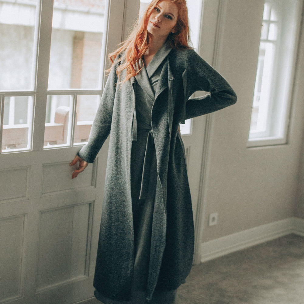 
                      
                        Woman in a long coat standing in a room with large windows.
                      
                    
