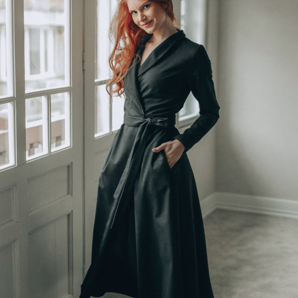 
                      
                        Woman in a wool wrap dress standing by a window in a room with light-colored walls.
                      
                    