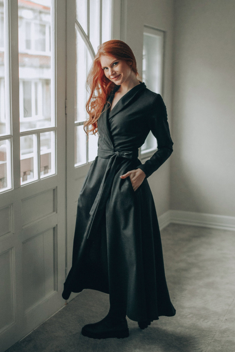 Woman wearing elegant black wool wrap dress with shawl collar and side pockets indoors by window