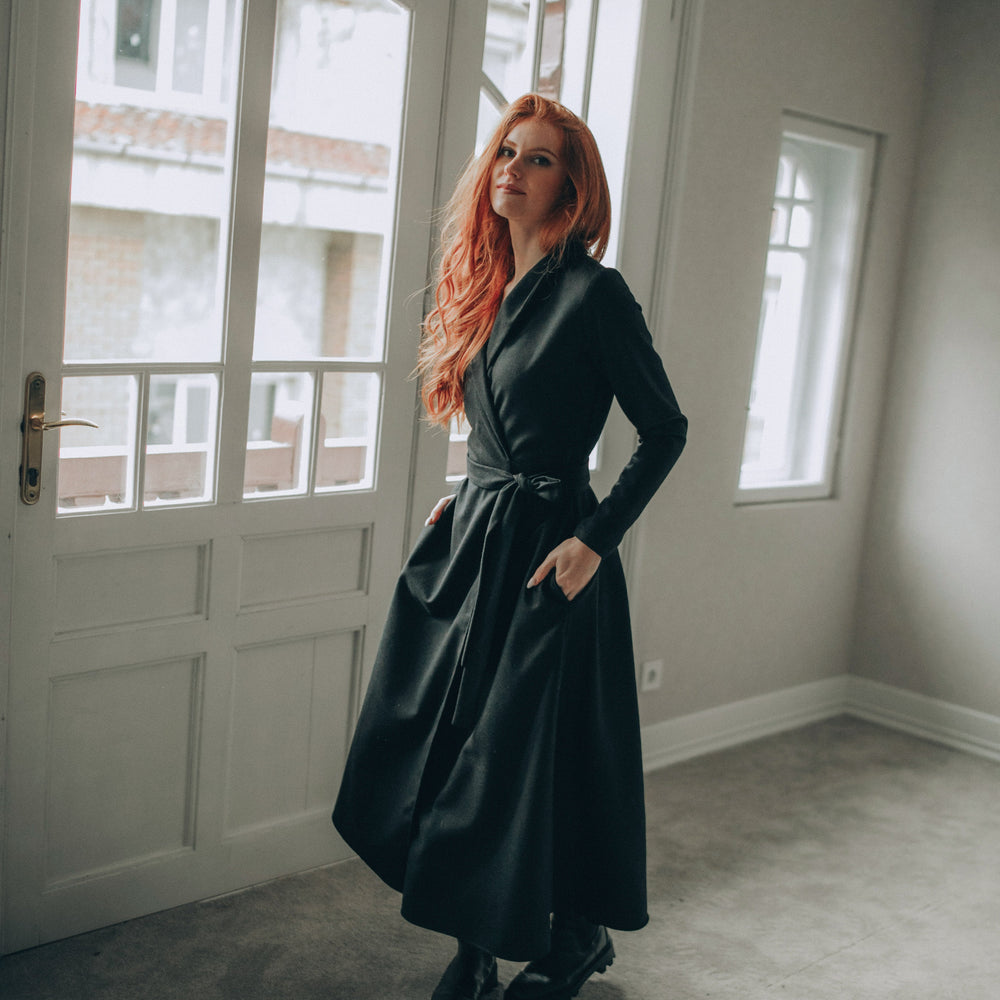 
                      
                        Woman in a wool wrap dress standing by a window in a room with light-colored walls.
                      
                    