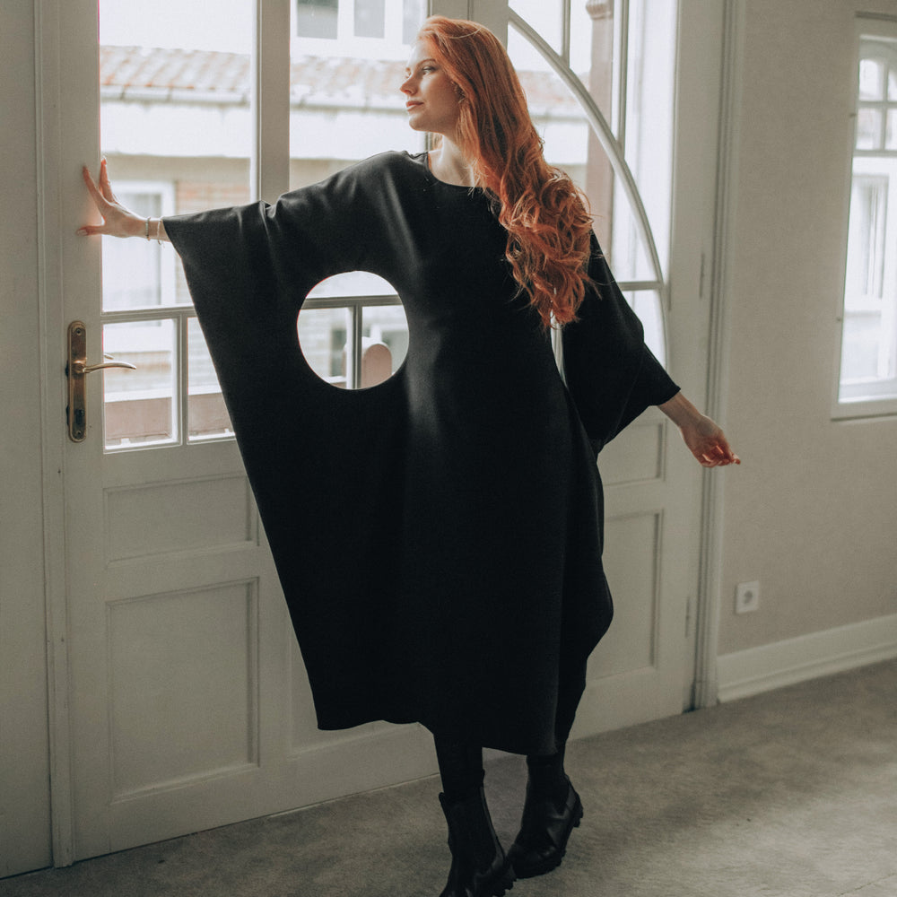 Woman wearing a black midi dress with a circular cutout in a room with large windows.