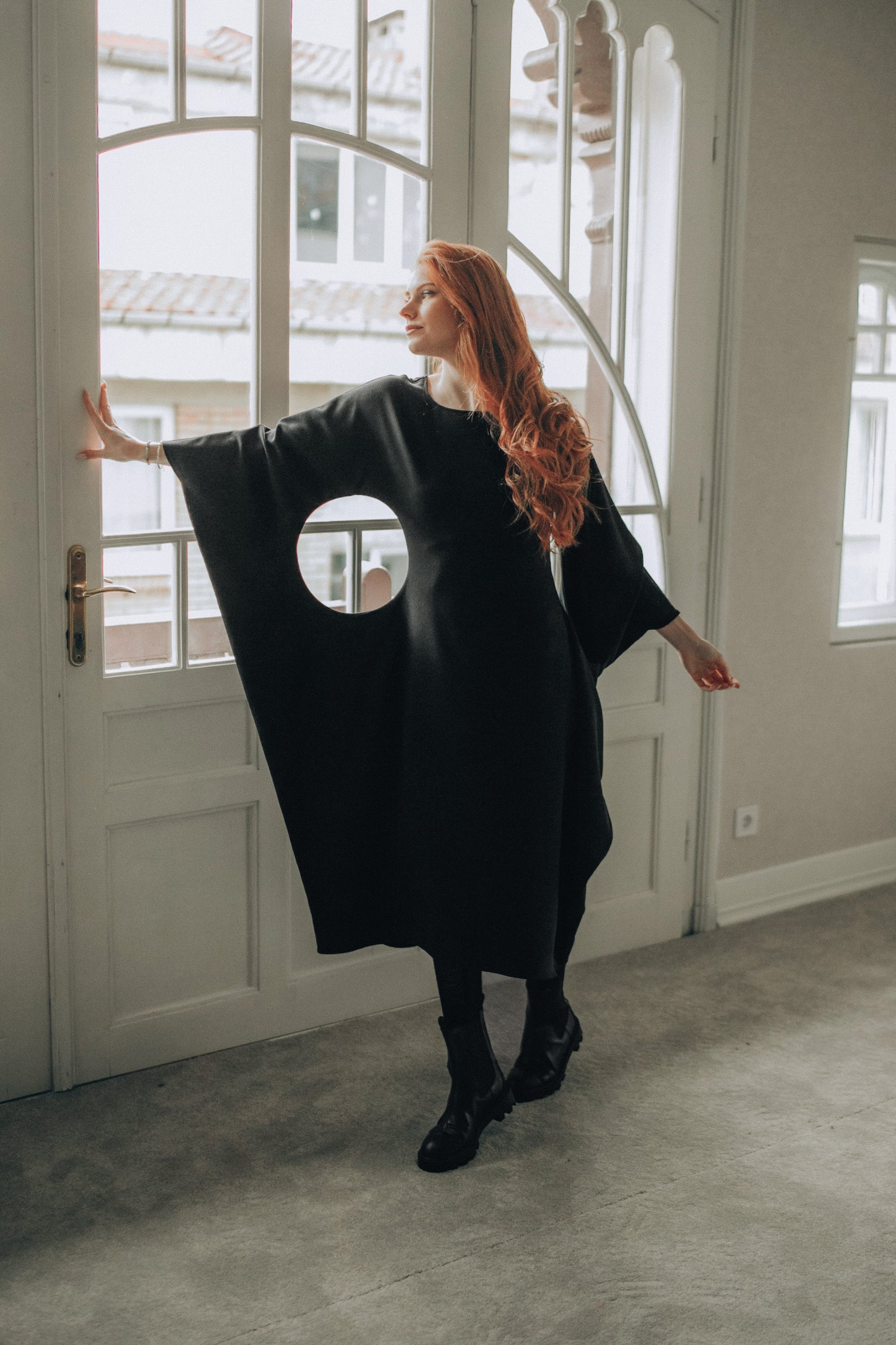 Woman wearing a black midi dress with a circular cutout in a room with large windows.