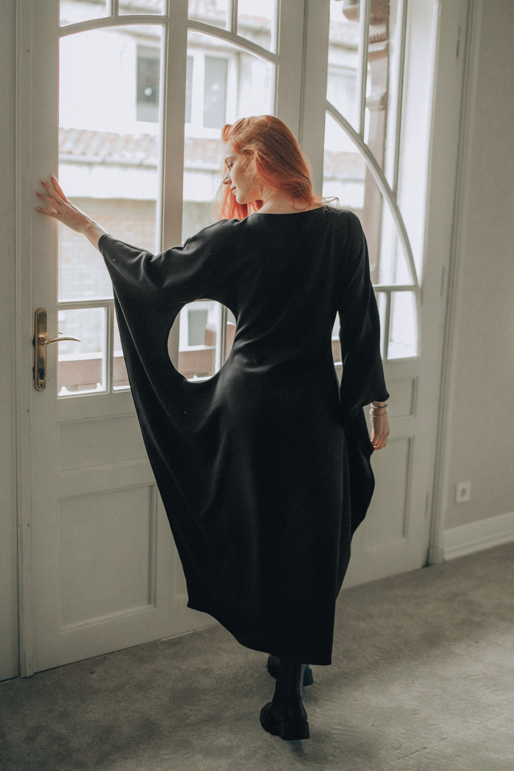 Woman wearing black batwing sleeve midi dress standing by a window