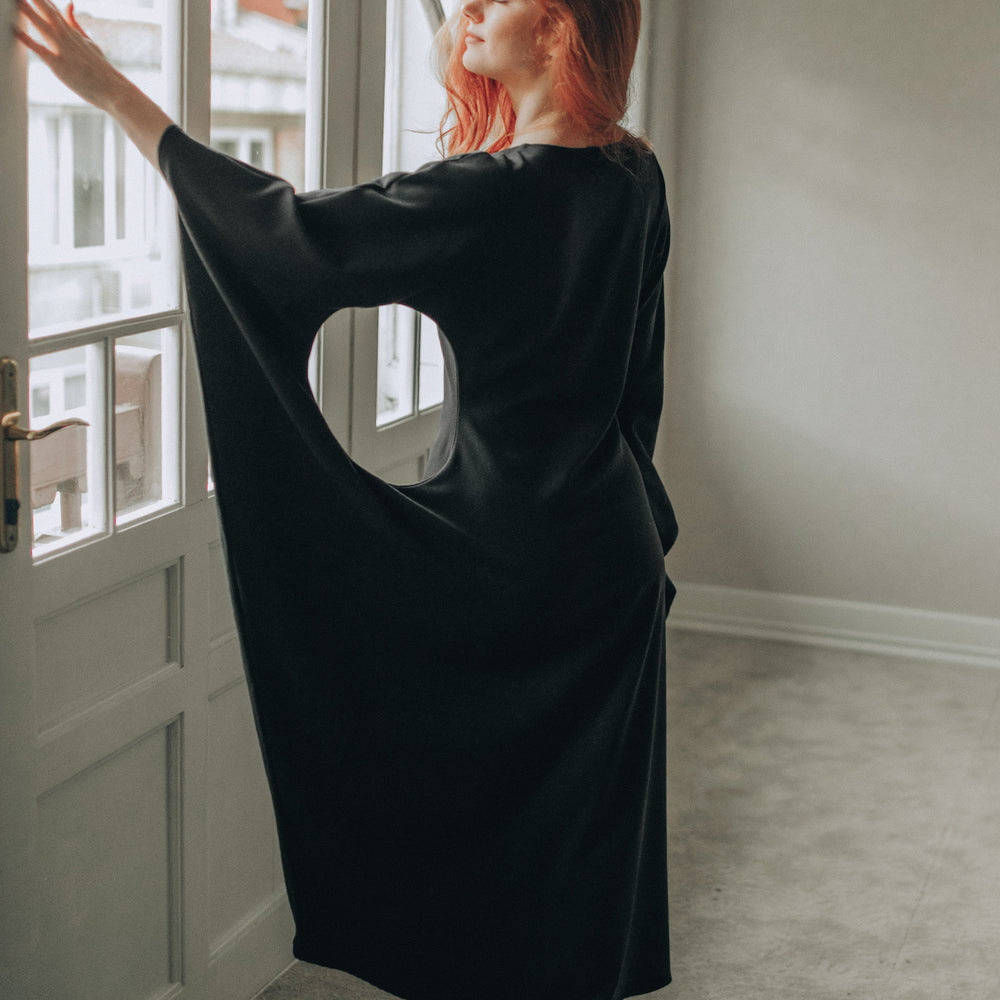 
                      
                        Person wearing a black dress standing by a window in a room with light-colored walls and carpet.
                      
                    
