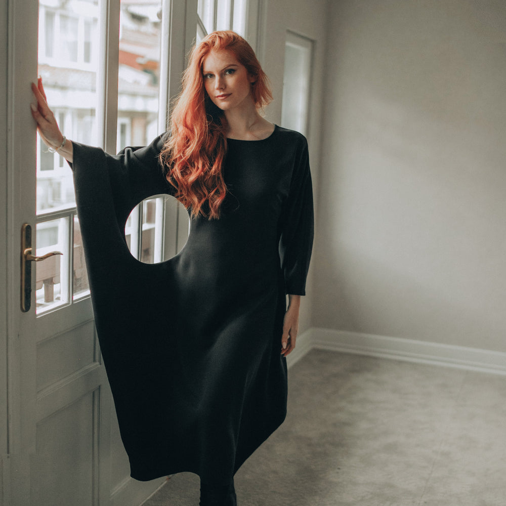 
                      
                        Woman in a black dress standing in a room with large windows.
                      
                    