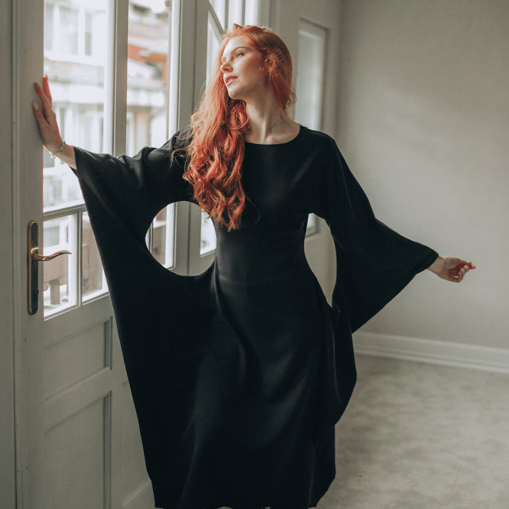 
                      
                        Woman in a black dress with wide sleeves standing in a room with large windows.
                      
                    