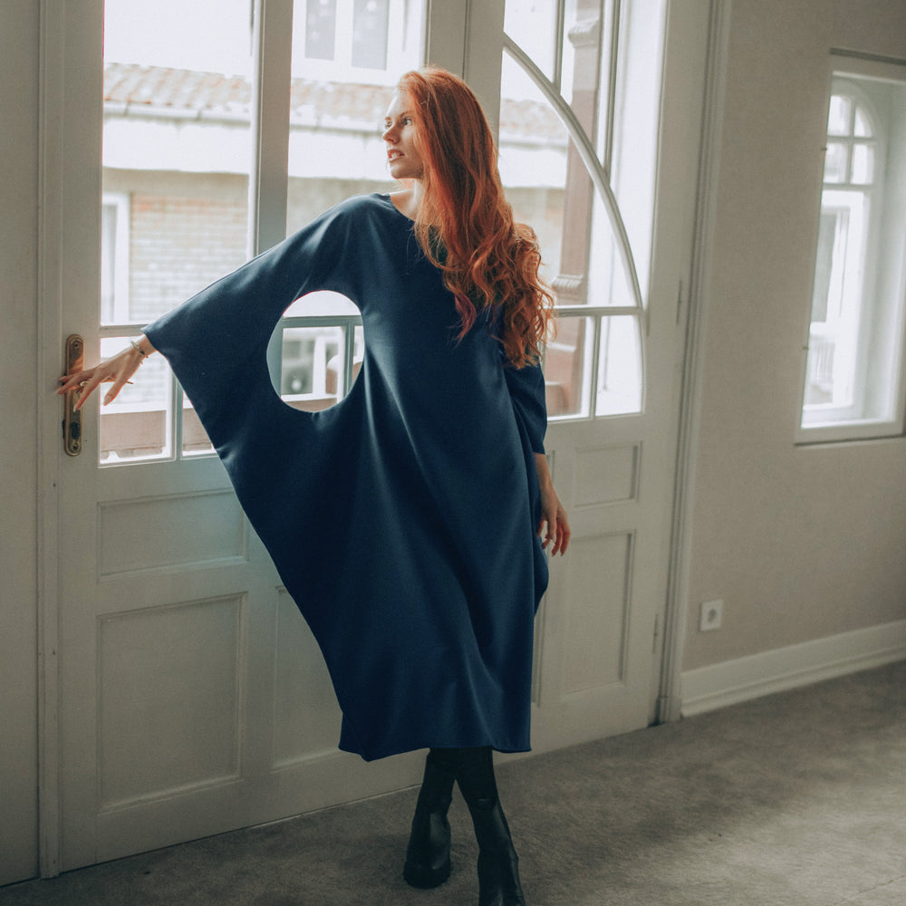
                      
                        Woman in a petrol blue dress standing in a room with large windows.
                      
                    