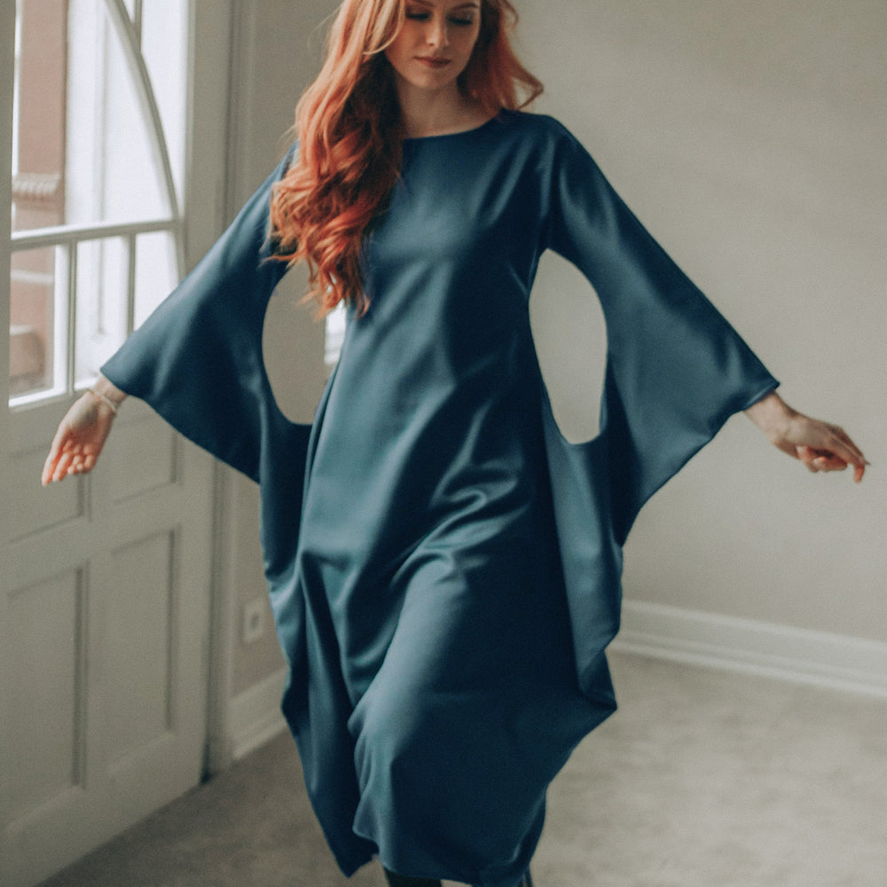 
                      
                        Woman in a petrol blue midi dress with wide sleeves standing in a room with a neutral color scheme.
                      
                    