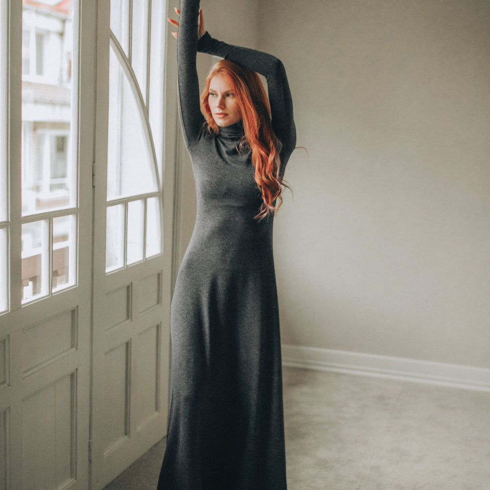 Woman in a long dark gray dress standing in a room with large windows.