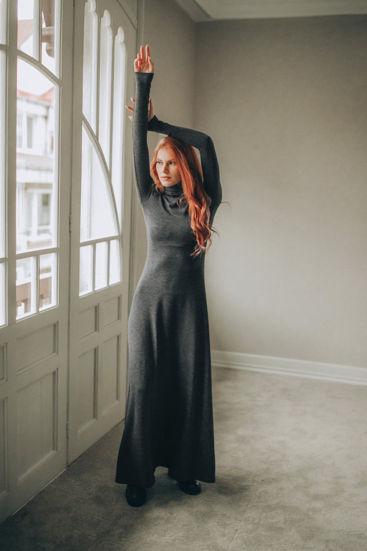 Woman in a long dark gray dress standing in a room with large windows.