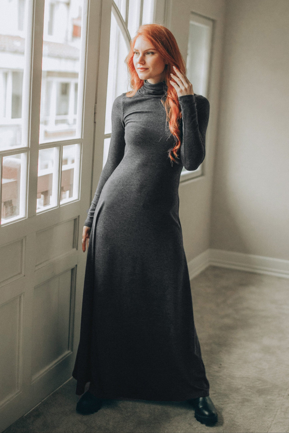 Woman wearing a fitted charcoal cotton maxi turtleneck dress with long sleeves near window