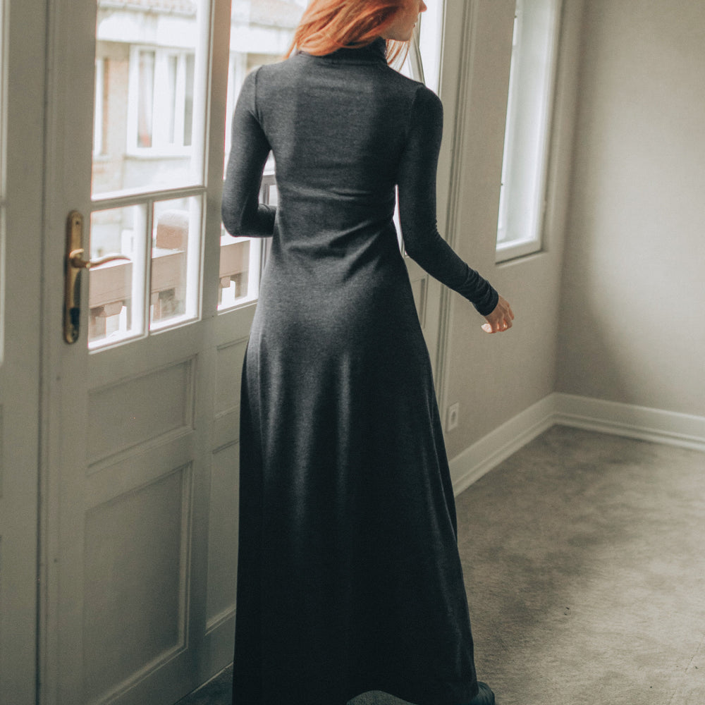 
                      
                        Person in a long dark dress standing in a room with a door and window.
                      
                    