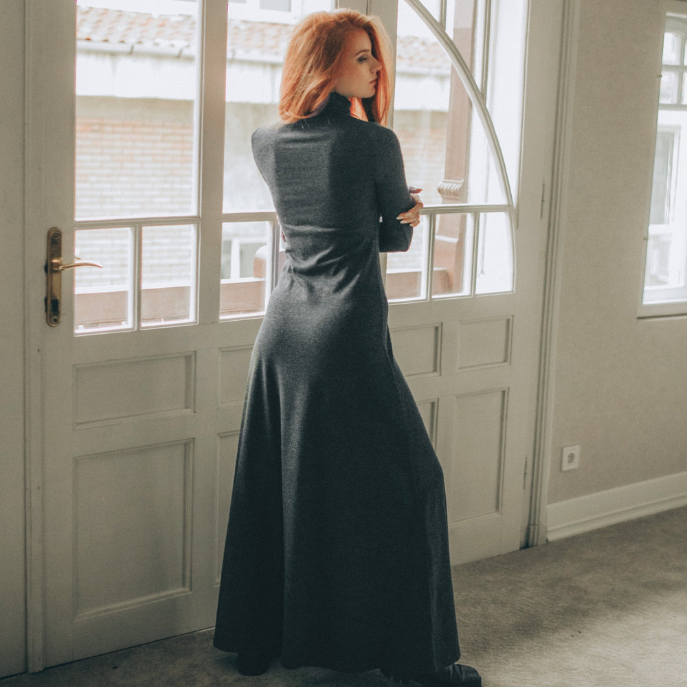 
                      
                        Woman in a long black dress standing in a room with large windows.
                      
                    