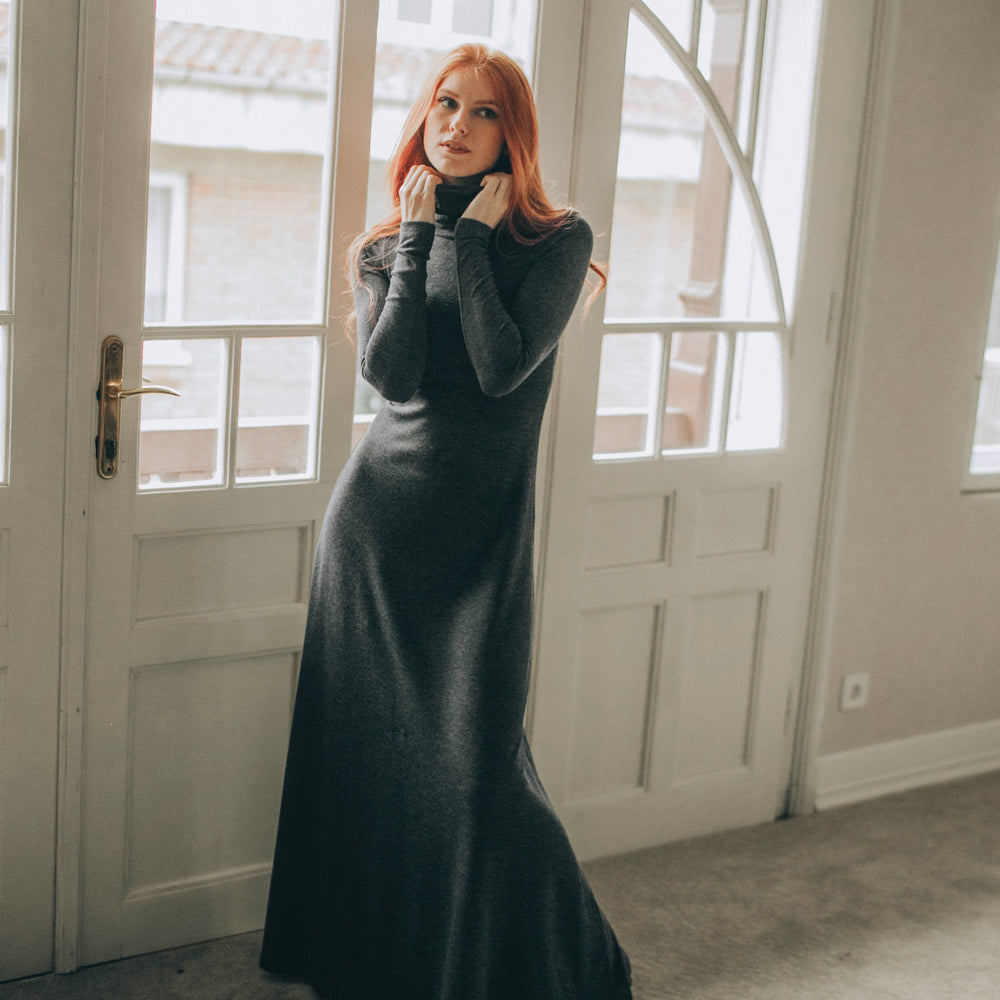 
                      
                        Woman in a long dark dress standing in a room with large windows.
                      
                    