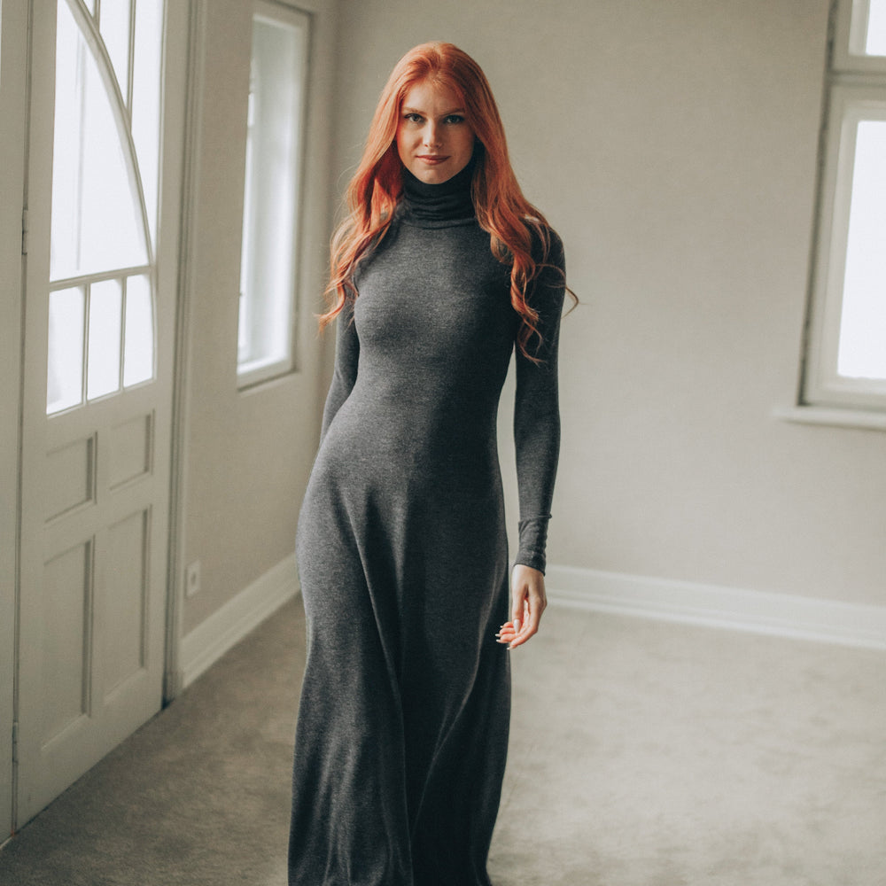 
                      
                        Woman in a long gray dress standing in a room with neutral walls and a door.
                      
                    