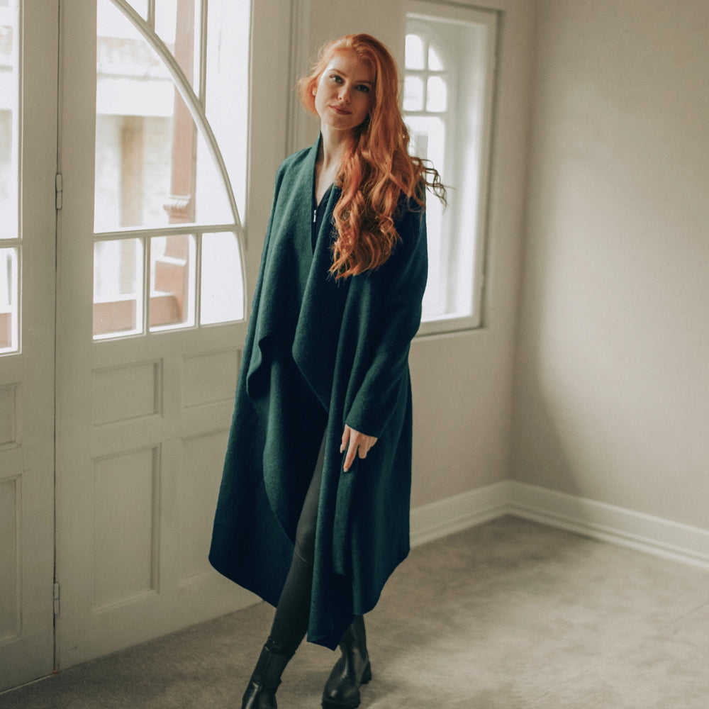 
                      
                        Woman in a green coat standing in a room with large windows.
                      
                    