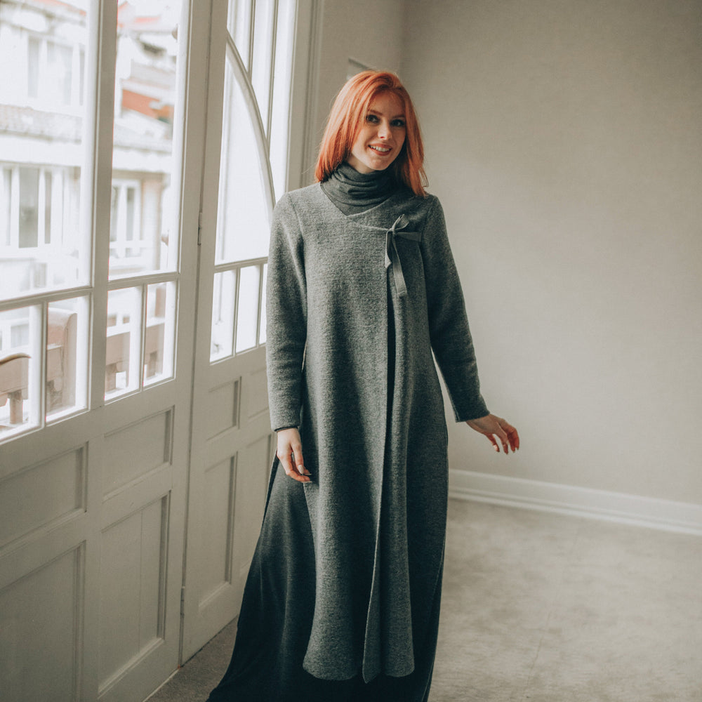 
                      
                        Woman in a long gray cardigan standing in a room with large windows.
                      
                    