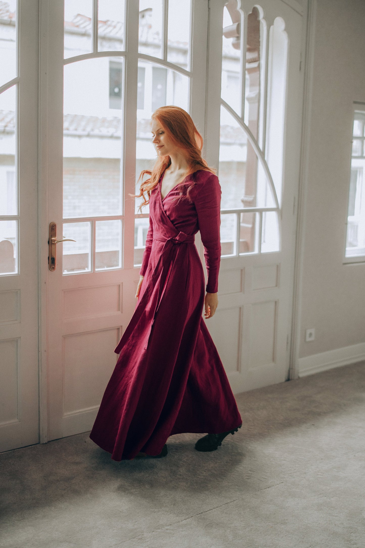 Woman in a burgundy linen dress standing in a room with large windows.