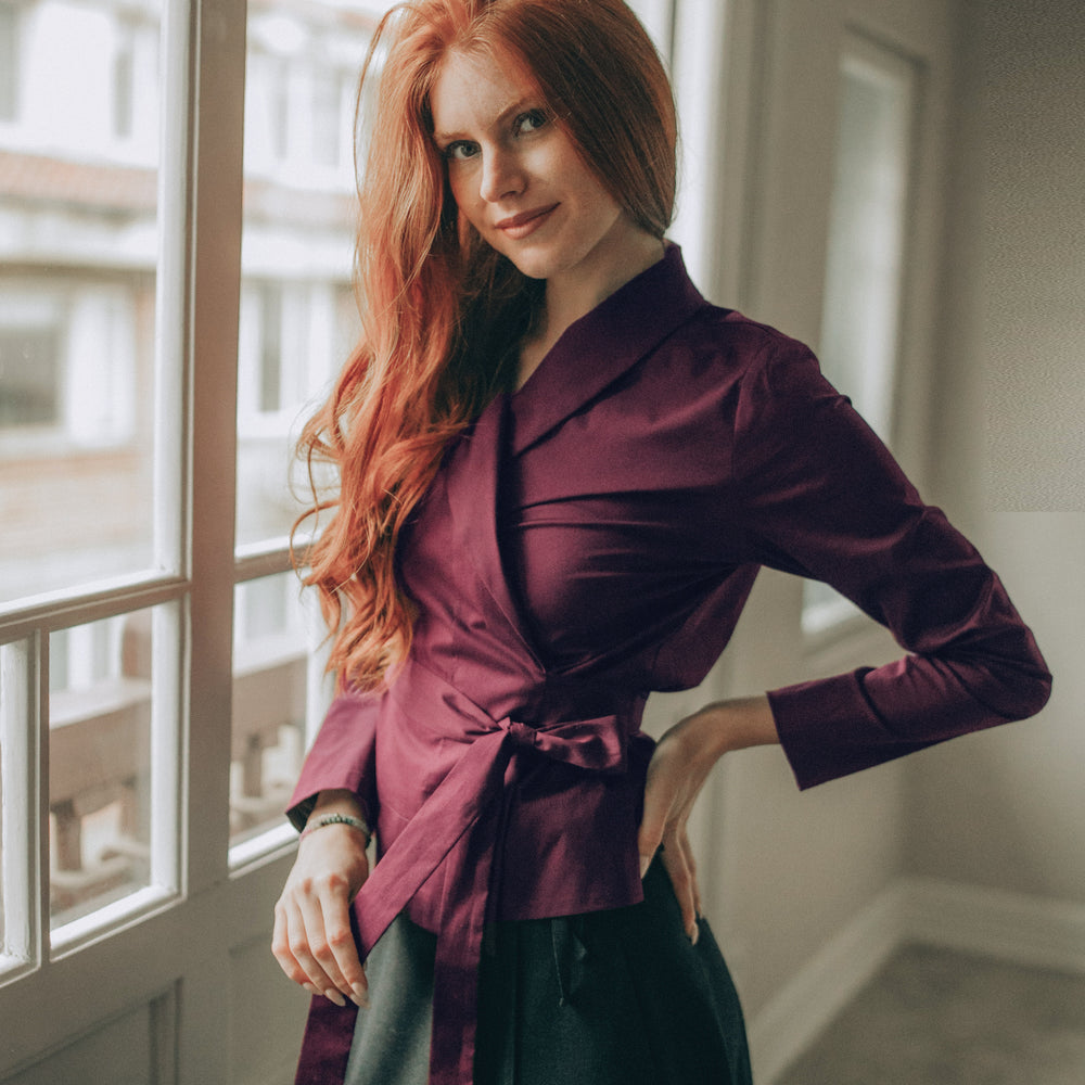 Woman in a burgundy wrap top and black skirt standing by a window 