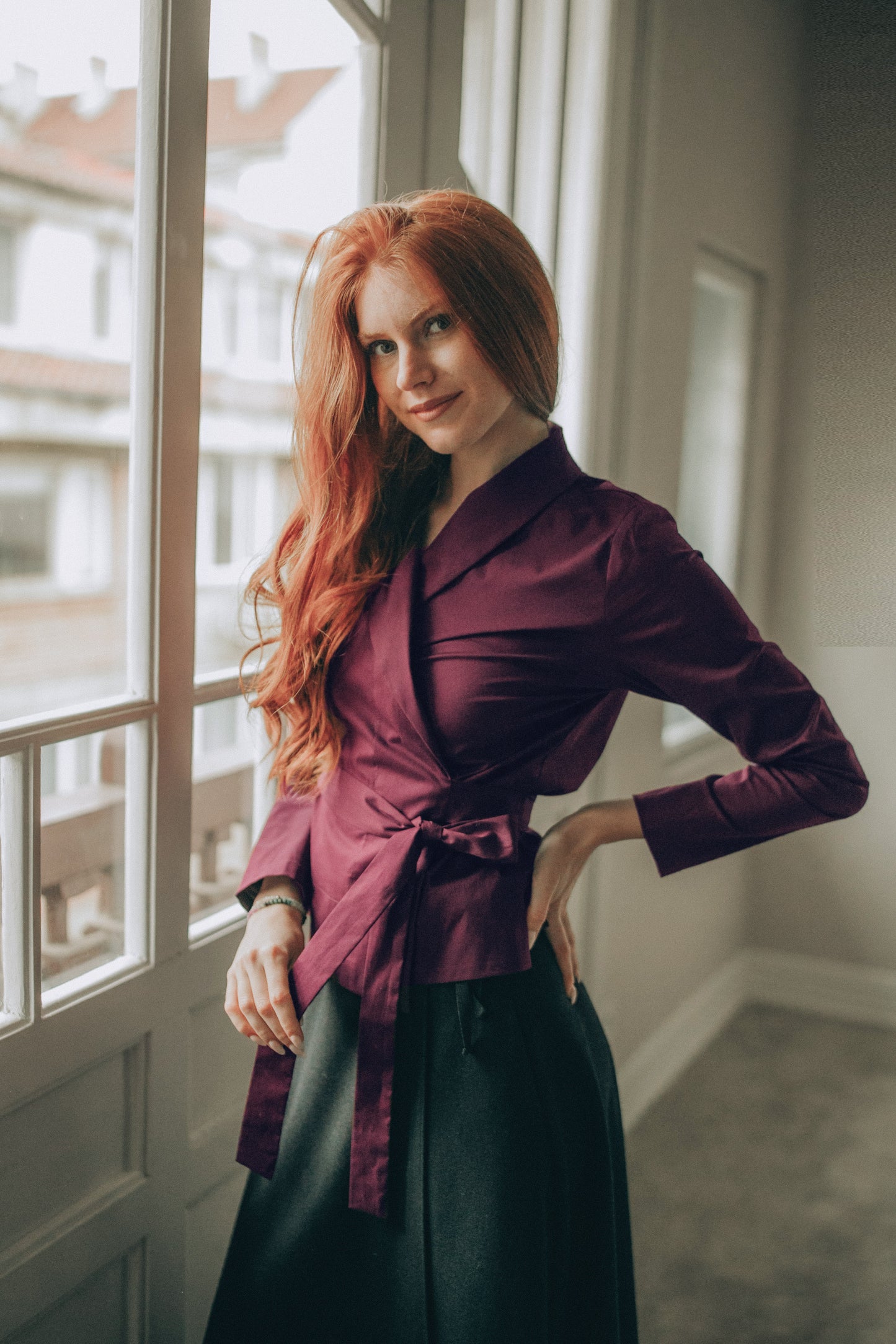 Woman in a burgundy wrap top and black skirt standing by a window 