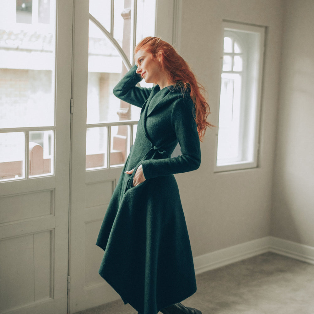 Woman in a dark green asymmetrical coat standing in a room with large windows.