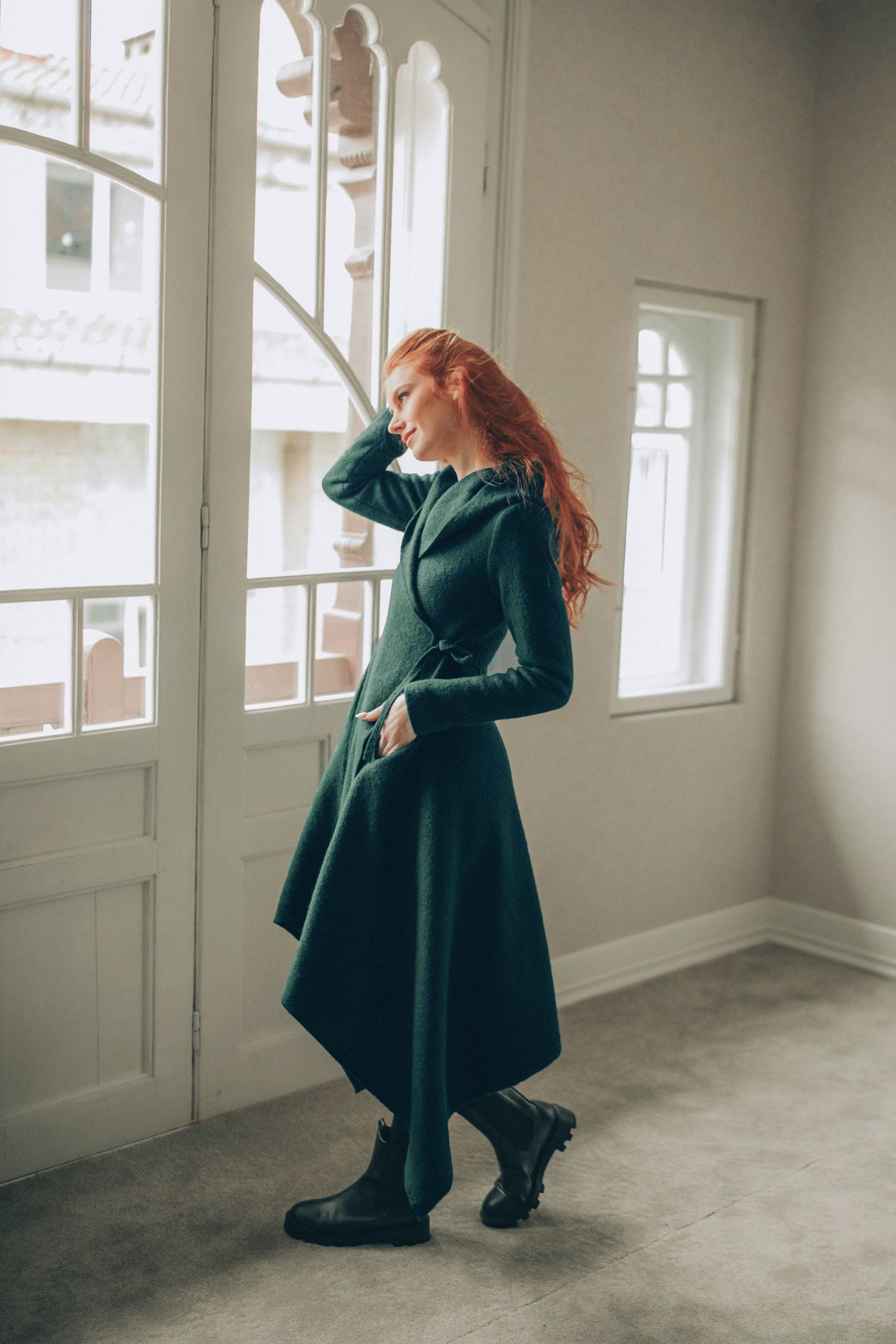 Woman in a dark green asymmetrical coat standing in a room with large windows.