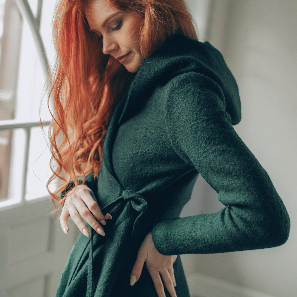 
                      
                        Woman in a dark green asymmetrical coat standing in a room with large windows.
                      
                    