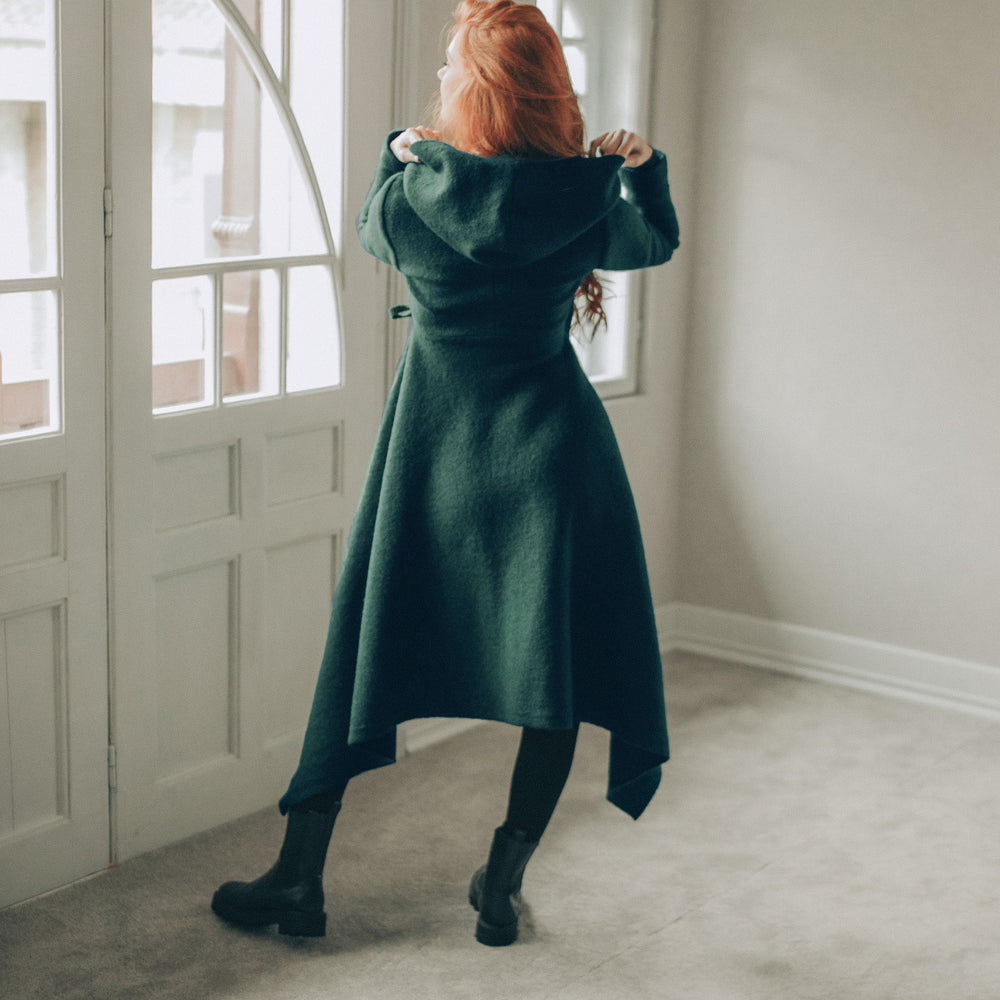 
                      
                        Woman in a dark green asymmetrical coat standing in a room with large windows.
                      
                    