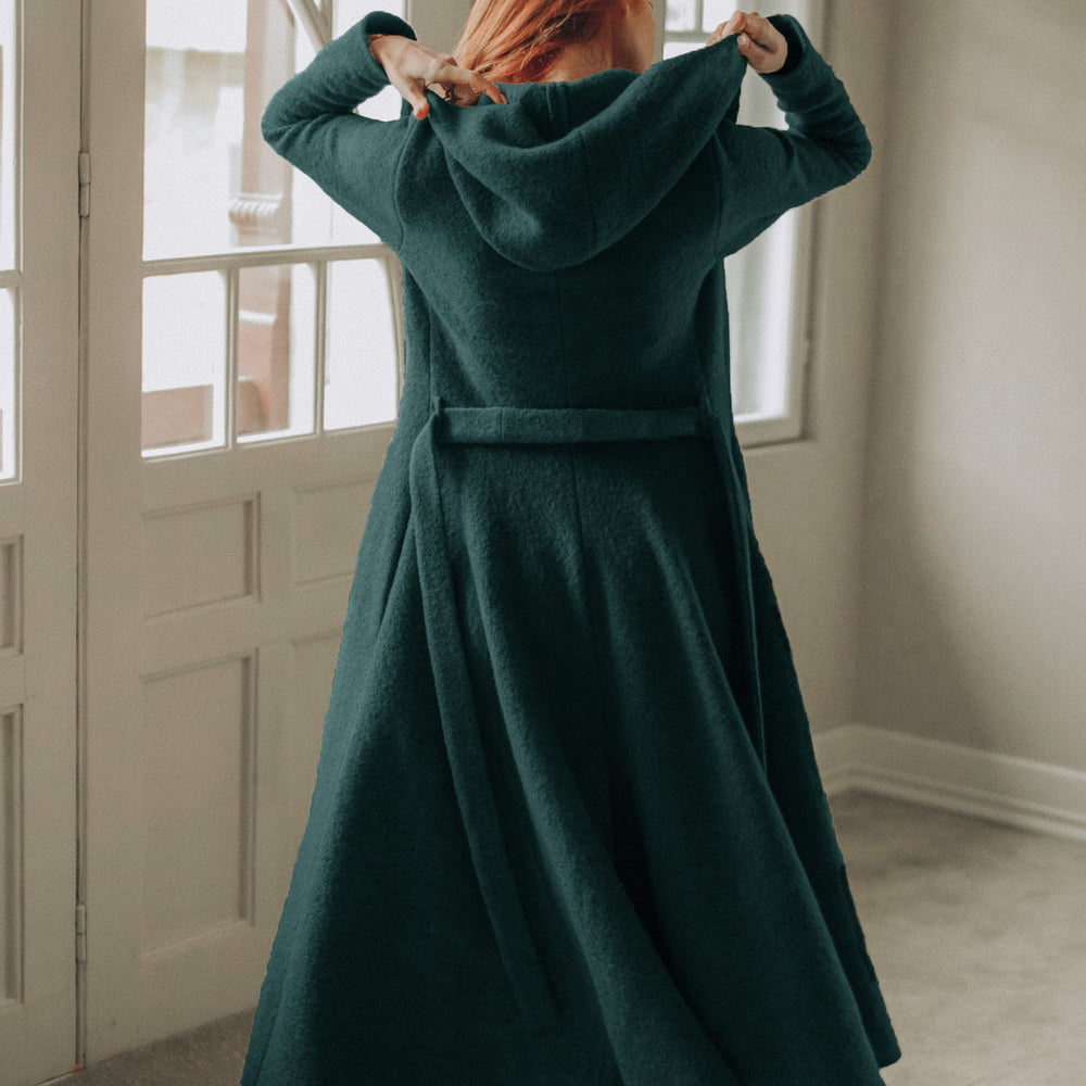 Back view of a woman wearing a dark green maxi wool hooded cardigan coat with an adjustable belt and flowing skirt