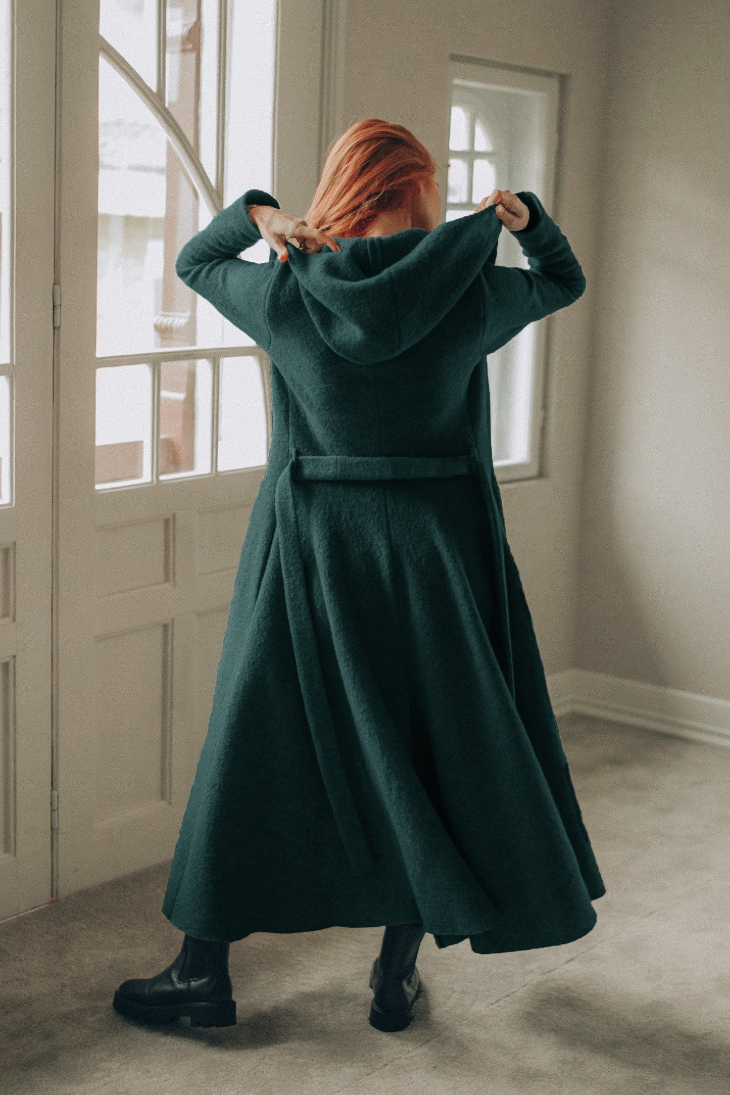 Back view of a woman wearing a dark green maxi wool hooded cardigan coat with an adjustable belt and flowing skirt