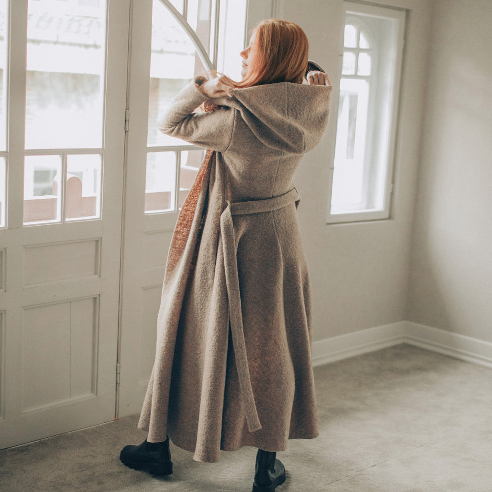Woman in a long beige coat standing in a room with large windows.