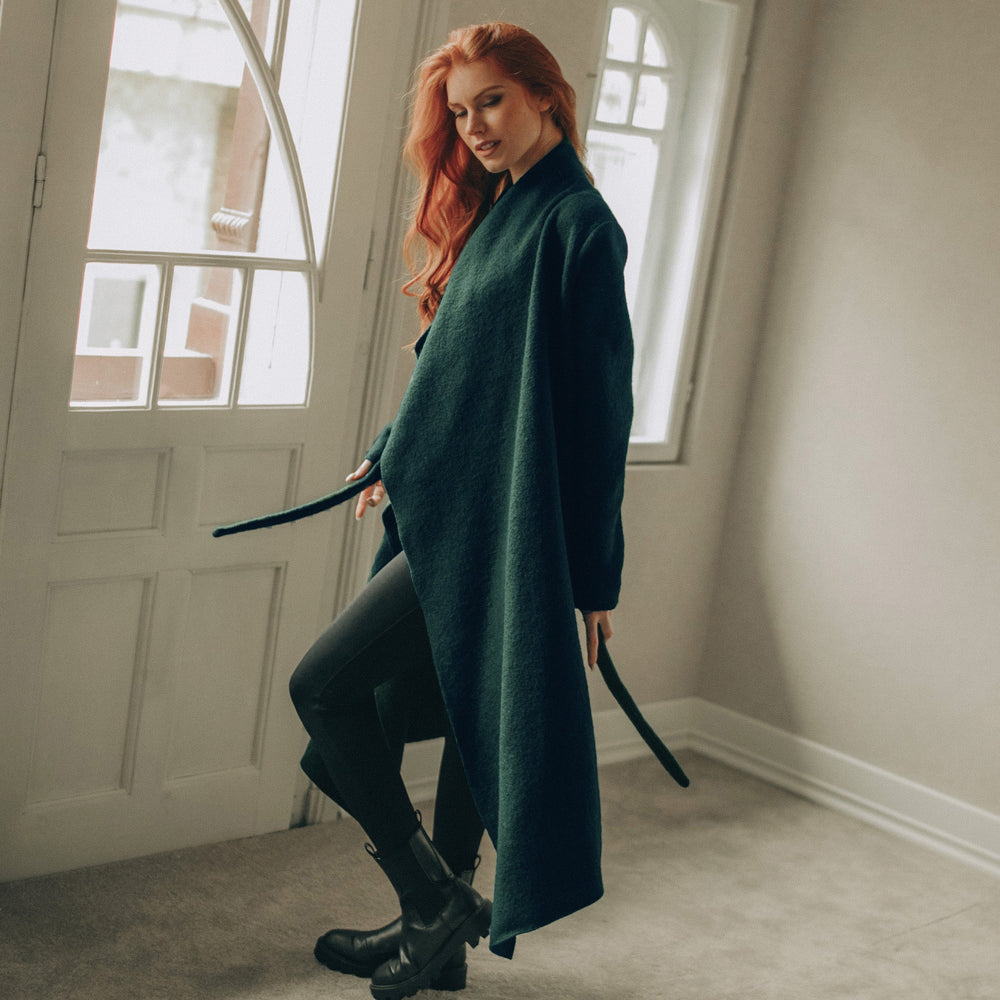 
                      
                        Woman in a dark green coat standing in a room with large windows.
                      
                    
