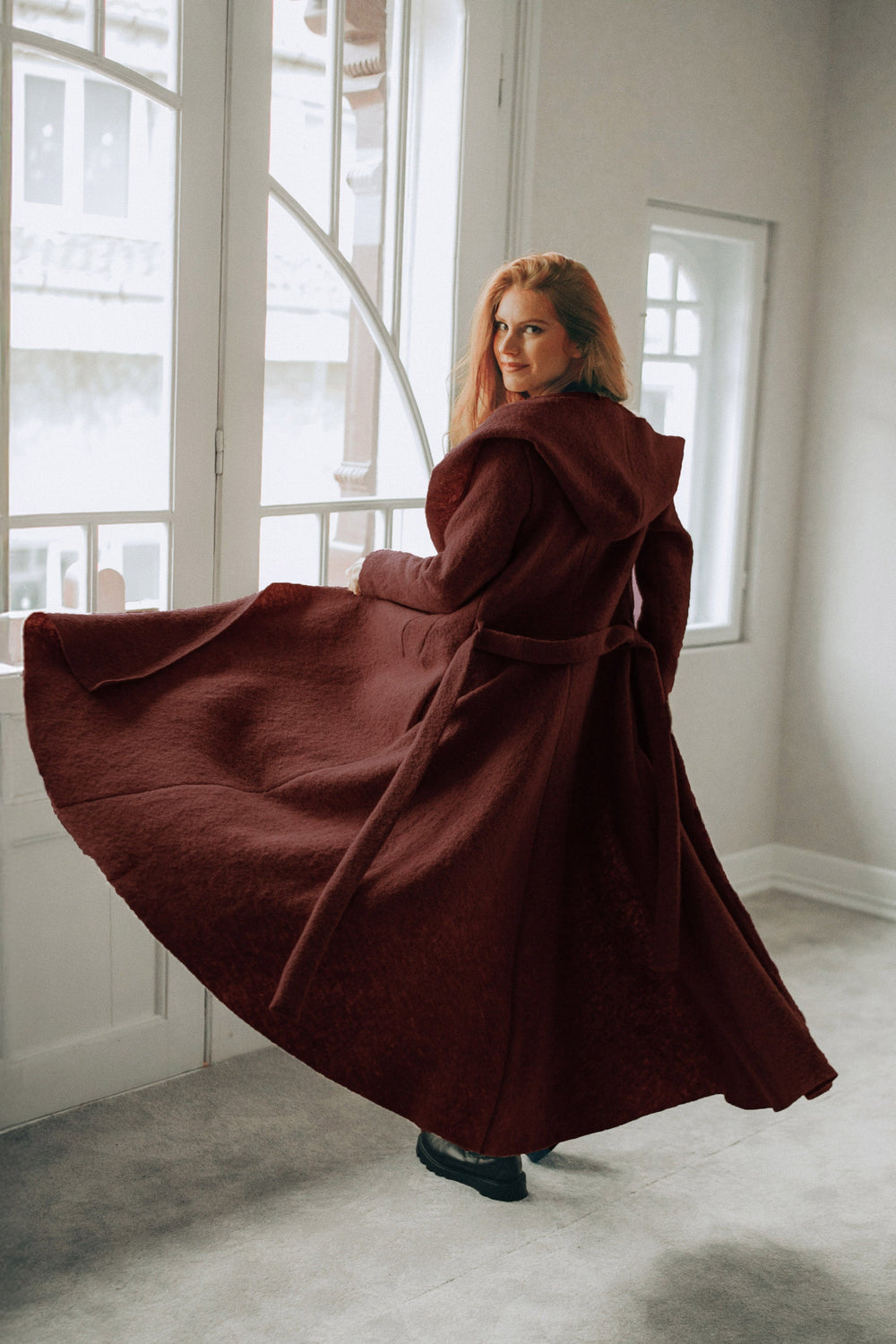 Woman wearing a flowing burgundy maxi wool hooded cardigan coat with an adjustable belt and hood indoors