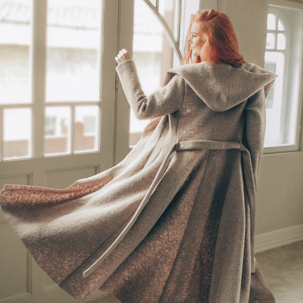 Maxi wool hooded cardigan coat with princess silhouette and adjustable belt, flowing long skirt, cozy hood, and deep pockets, worn indoors.
