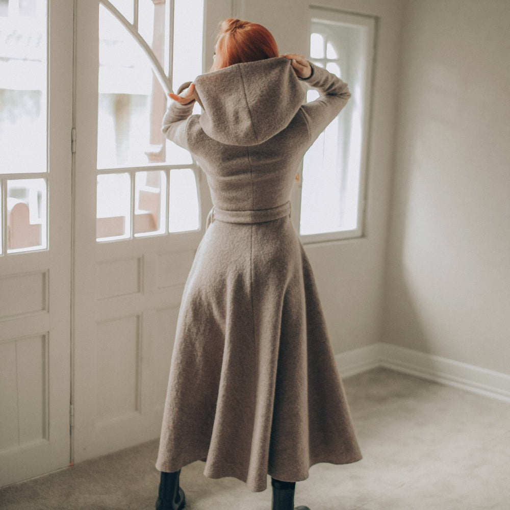 
                      
                        Woman wearing a long beige coat standing in a room with large windows.
                      
                    
