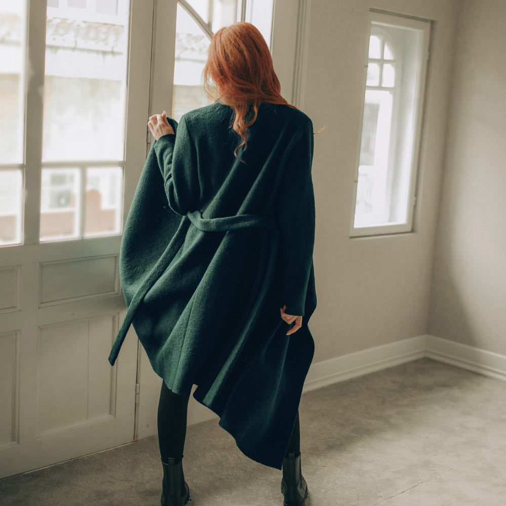 
                      
                        Woman in a green coat standing in a room with large windows.
                      
                    