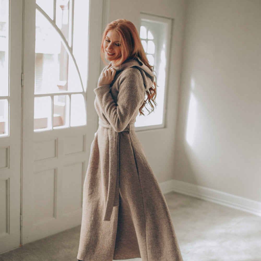 
                      
                        Woman in a long beige coat standing in a room with large windows.
                      
                    
