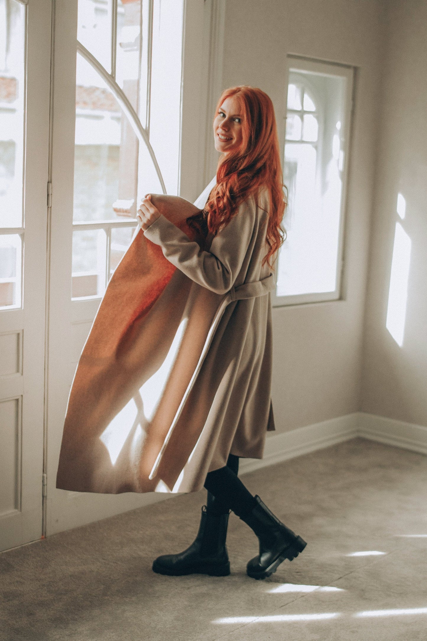 Woman with red hair standing in a sunlit room holding a coat