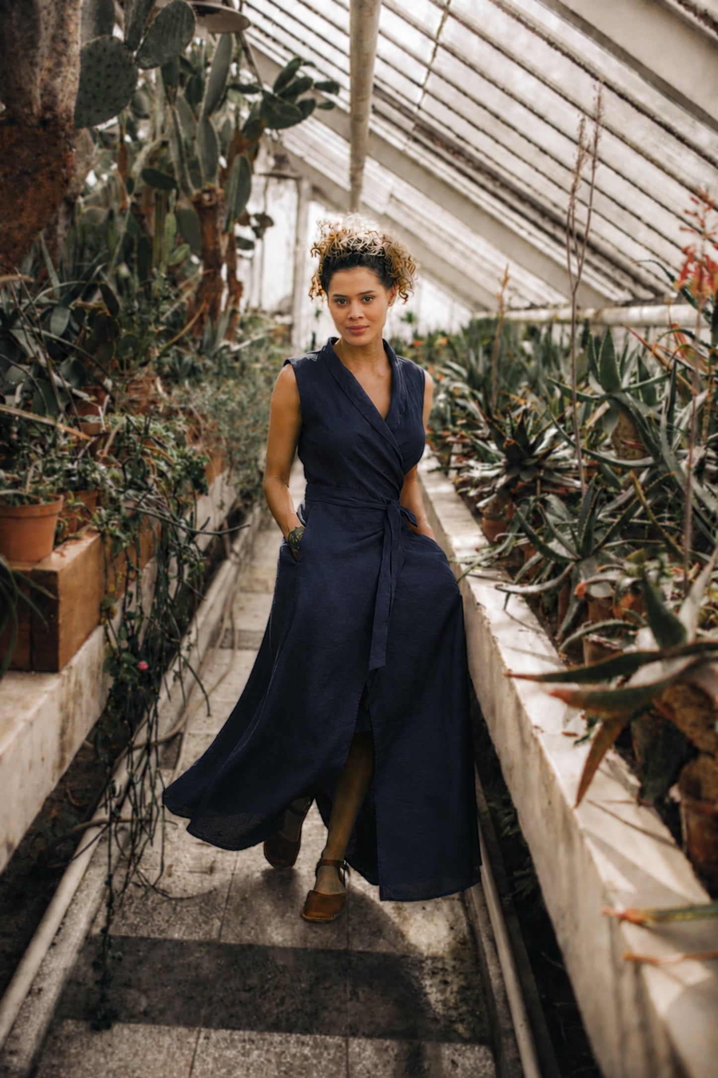 Woman in a navy blue linen maxi dress standing in a greenhouse filled with plants.
