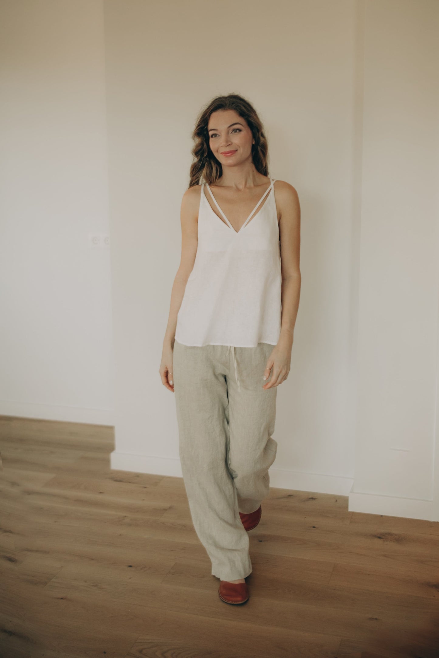 Woman wearing a white linen  top and beige pants standing in a room with wooden floor and white walls.